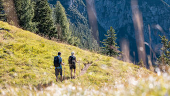 Two hikers walking along a grassy mountain trail in Alpbach, Austria, with alpine peaks in the background and wildflowers lining the path.