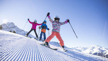 A family build up speed as they run down a slope together, all brightly dressed and surrounded by vast, snowy peaks.
