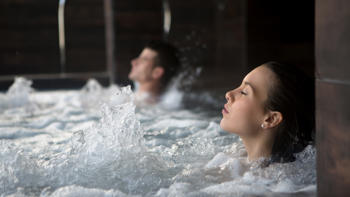 Two people relax side by side in a steaming hot tub, surrounded by calming ambiance and soft, warm light.