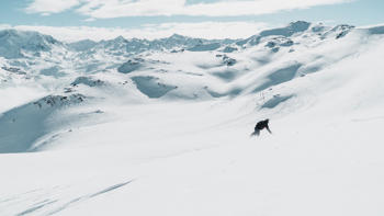 A lone skier kicks up powered snow as they rush down the side of a white mountain, surrounded by white peaks and valleys.