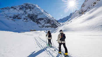Two skiers begin their climb of a nearby mountain, taking in the surrounding valleys and peaks under a bright blue sky.