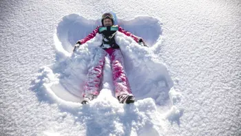 A girl wearing colourful ski gear creates a snow angel on the ground, having fun under a bright sun.