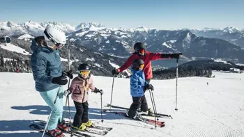 Two parents teach their children to ski, encouraging them down a gentle slope amidst the surrounding mountains and valleys.