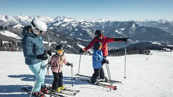 Two parents teach their children to ski, encouraging them down a gentle slope amidst the surrounding mountains and valleys.