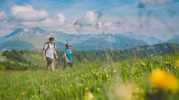 Couple walking through a green meadow in Morzine, surrounded by wildflowers and distant mountains.