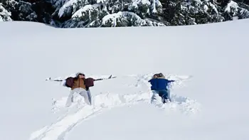 Two skiers make snow angels on the ground, enjoying themselves as they look up at a bright sun and clear skies.