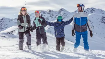A family dressed in full ski gear playfully kick up powdered snow as they relax at the peak of a white mountain.