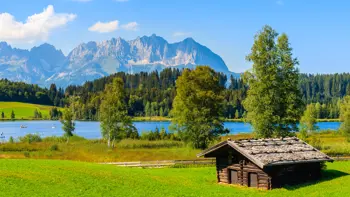 View of Lake Schwarzsee near Kitzbühel with a wooden hut in the foreground, calm water reflecting surrounding trees and alpine peaks in the distance.