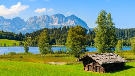 View of Lake Schwarzsee near Kitzbühel with a wooden hut in the foreground, calm water reflecting surrounding trees and alpine peaks in the distance.