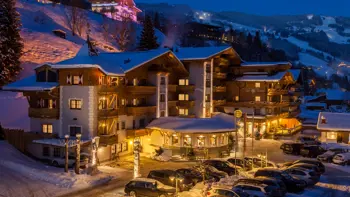 Hotel Die Sonne in Saalbach shines warmly against the snowy winter landscape, its elegant alpine facade adorned with wooden balconies and soft lighting, nestled among snow-covered trees with the Austrian Alps providing a breathtaking mountain backdrop.