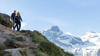 Walkers stroll along a scenic alpine trail in Saas Fee, surrounded by towering snow-capped peaks under a clear blue sky.