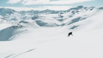 A lone skier kicks up powered snow as they rush down the side of a white mountain, surrounded by white peaks and valleys.