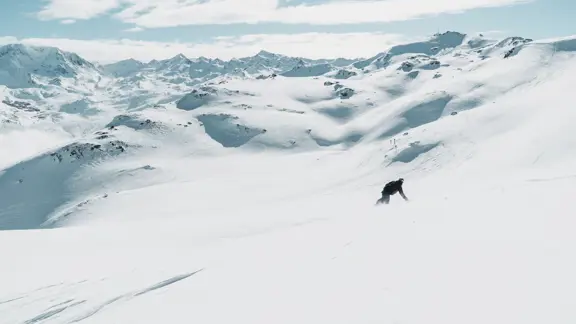 A lone skier kicks up powered snow as they rush down the side of a white mountain, surrounded by white peaks and valleys.