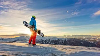 A lone skier stands at the top of a slope, looking out across the snowy landscape under a bright blue sky.