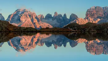 A serene lake reflects the surrounding trees and sky, with the majestic peaks of the Madonna di Campiglio Dolomites rising dramatically in the background.