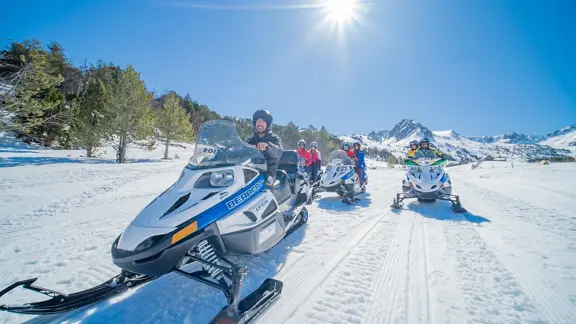 A group rush through the Andorra mountains on snowmobiles, with a bright blue sky and brilliant sun shining down on them as they go.