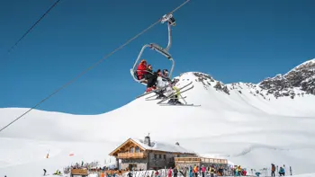 A family rides a ski lift up a snowy mountain slope in Val d’Isère, bundled in winter gear and smiling, with panoramic views of rugged peaks and alpine terrain