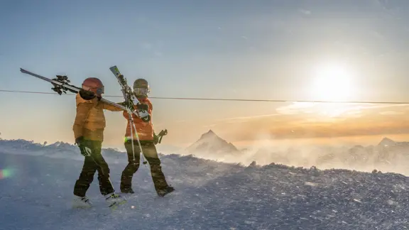 A couple of skiers walk along the top of a snowy peak, watching the sunset as they take in the beautiful views of mountains and valleys.