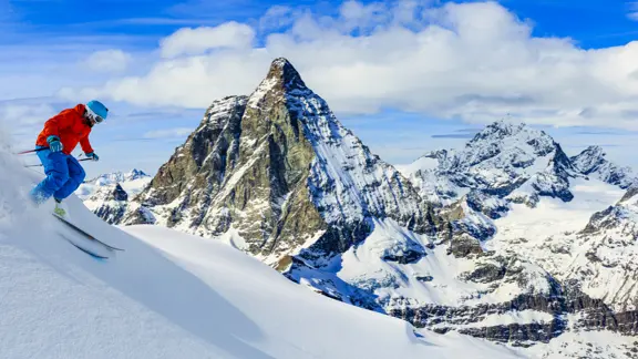 A lone skier rushes down a piste in Zermatt, kicking up powdered snow as they take in the nearby mountains and snowy valleys.