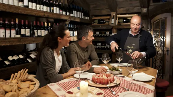 A couple being poured a glass of wine at the Hotel's restaurant, with several rows of bottles shelved behind them.