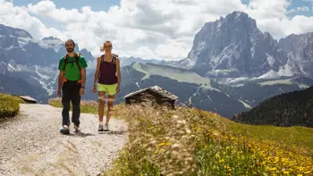 A couple walks along a scenic trail in the Dolomite mountains near Selva, with towering limestone peaks and alpine meadows in the background.