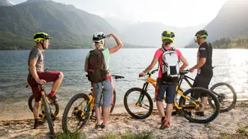 Four cyclists take a break beside Lake Bohinj, resting with their bikes near the calm water, surrounded by mountains and greenery