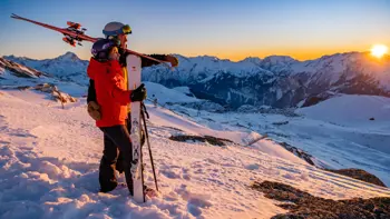 A pair of skiers watch the sunset go down together, holding one another as they look out across vast snowy landscapes and mountains.