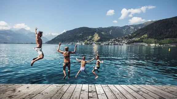 Family jumping into the clear water beside Lake Zell am See on a sunny day, surrounded by mountains.