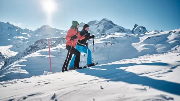 A pair of skiers enjoy snowshoeing up a mountainside, taking in the views of endless snowy peaks and clear blue skies.