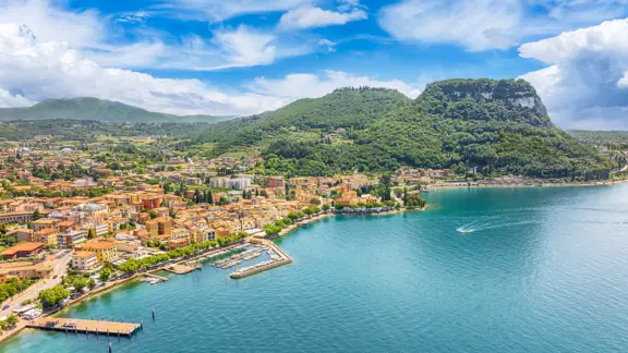 Panoramic view of Lake Garda’s lakeside in Garda town, featuring calm blue waters, boats docked along the shore, and surrounding hills under a bright sky.