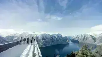 A group stare over a view point in Myrkdalen, looking at a vast landscape of snowy mountains, lush fields and wide, flowing rivers all under a bright blue sky.
