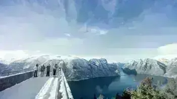 A group stare over a view point in Myrkdalen, looking at a vast landscape of snowy mountains, lush fields and wide, flowing rivers all under a bright blue sky.