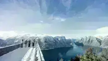 A group stare over a view point in Myrkdalen, looking at a vast landscape of snowy mountains, lush fields and wide, flowing rivers all under a bright blue sky.
