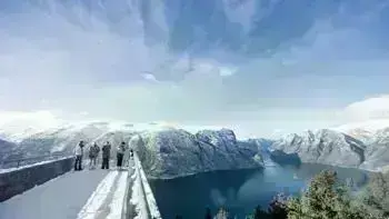 A group stare over a view point in Myrkdalen, looking at a vast landscape of snowy mountains, lush fields and wide, flowing rivers all under a bright blue sky.