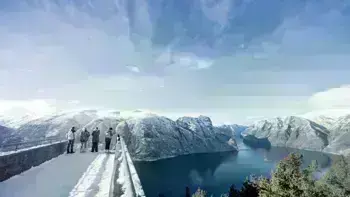 A group stare over a view point in Myrkdalen, looking at a vast landscape of snowy mountains, lush fields and wide, flowing rivers all under a bright blue sky.