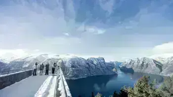 A group stare over a view point in Myrkdalen, looking at a vast landscape of snowy mountains, lush fields and wide, flowing rivers all under a bright blue sky.