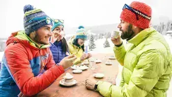 A group of skiers in colorful gear sit around an outdoor table, enjoying food and drinks with snowy mountains in the background.