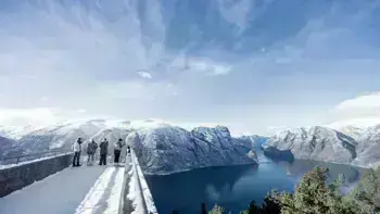 A group stare over a view point in Myrkdalen, looking at a vast landscape of snowy mountains, lush fields and wide, flowing rivers all under a bright blue sky.