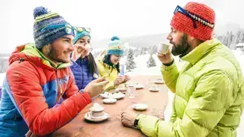 A group of skiers in colorful gear sit around an outdoor table, enjoying food and drinks with snowy mountains in the background.