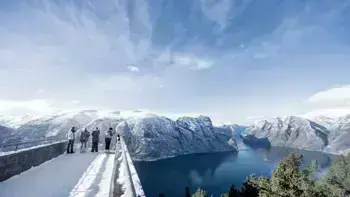 A group stare over a view point in Myrkdalen, looking at a vast landscape of snowy mountains, lush fields and wide, flowing rivers all under a bright blue sky.