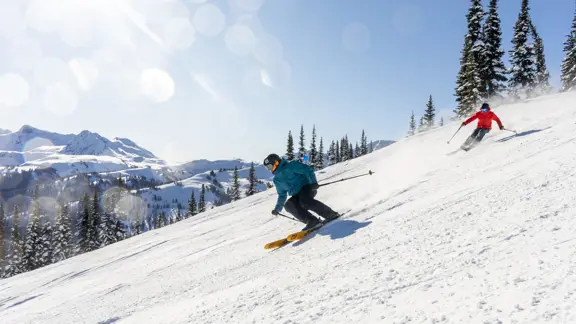 A pair of skiers rush down the slope, surrounded by views of endless mountains under a clear blue sky.