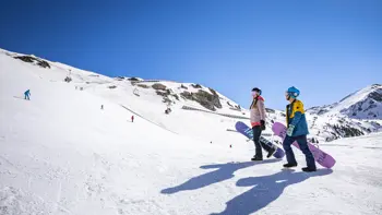 Two snowboarders climb a snowy slope in Austria, with a backdrop of several skiers rushing down the mountains under a bright blue sky.
