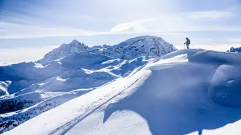 A lone skier stands at the top of a slope, looking out across the vast snowy landscape under a bright blue sky.