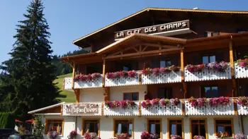 Hotel La Clef des Champs in Morzine presents a traditional alpine exterior, featuring a chalet-style wooden structure with balconies adorned with vibrant flower boxes, nestled amidst the picturesque mountain landscape