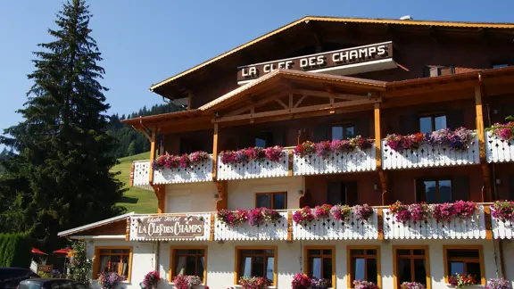Hotel La Clef des Champs in Morzine presents a traditional alpine exterior, featuring a chalet-style wooden structure with balconies adorned with vibrant flower boxes, nestled amidst the picturesque mountain landscape