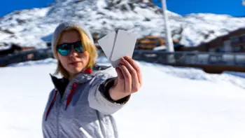 A woman holds her lift passes to the camera, with a background of snowy mountains under a bright blue sky.
