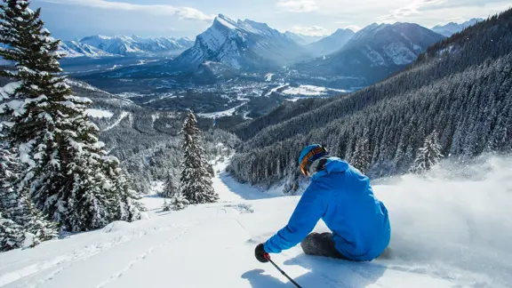A skier descends a snowy mountain with the stunning backdrop of Banff's mountains and valleys ahead of them.